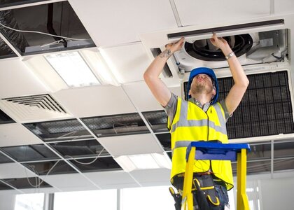 Image showing an electrician on a ladder fitting air conditioning unit into a ceiling. In a bright office an engineer in a hard hat stands on a ladder looking up as he fits an air conditioning unit into the ceiling.