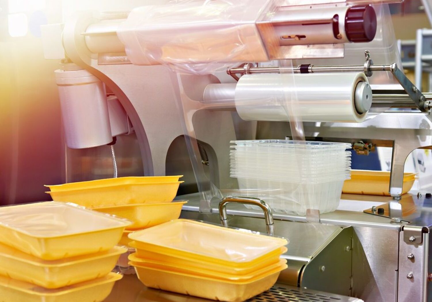 Image showing industrial equipment for food packaging in a factory. To the right an industrial machine is assembling yellow plastic food containers and wrapping them in transparent film. To the left sit three small stacks of the sealed packages.