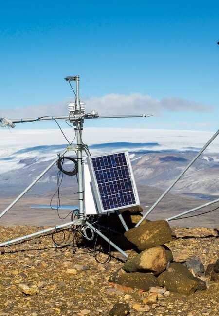 On a rocky ground in the mountains and framed by a blue sky sits a meteorological station. It is composed of a small monitor with a solar panel on the left and on the right a weathervane