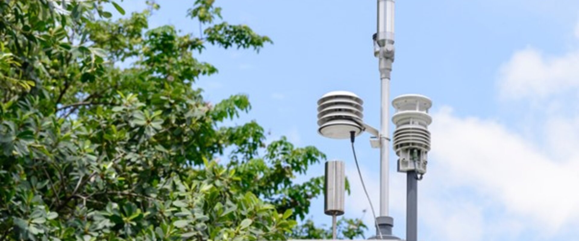 An air monitoring station In the centre of the frame, an air monitoring station is attached to a pole. It consists of a grey metal box with a handle pointing towards the viewer and three covered pylons, carrying sampling equipment, protruding from the top, as well as a small silver cylinder. In the background there is the canopy of a full, green tree, and a blue sky filled with soft clouds