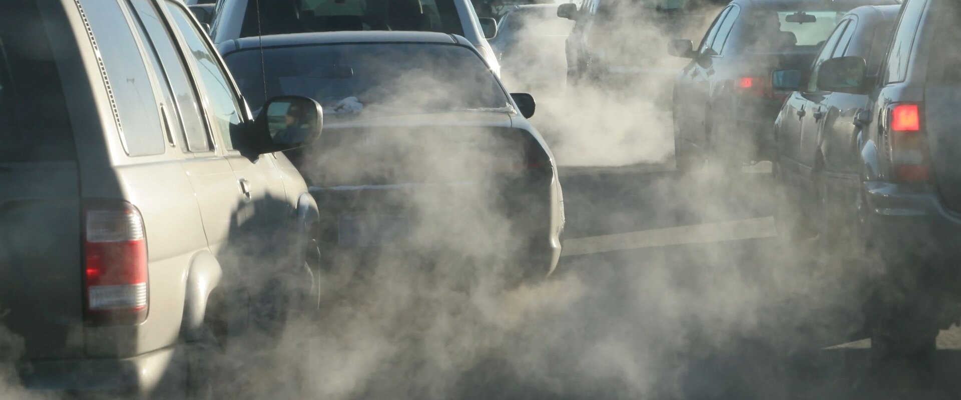 A photograph of a number of cars stopped at a traffic slight. The image is hazy with exhaust fumes coming from the vehicles’ exhausts