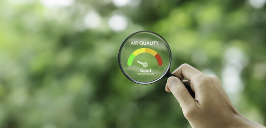 Against a blurry background of trees, in the foreground is a hand holding a magnifying glass with the words “Air quality – Good” in its centre